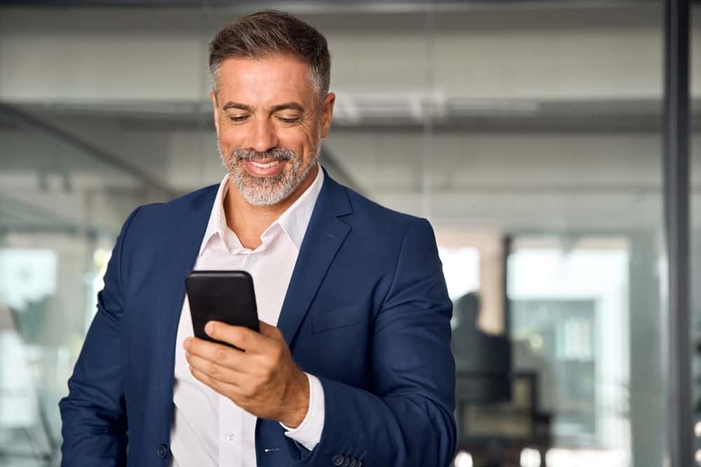 Smiling Mature Latin Or Indian Businessman Holding Smartphone In Office